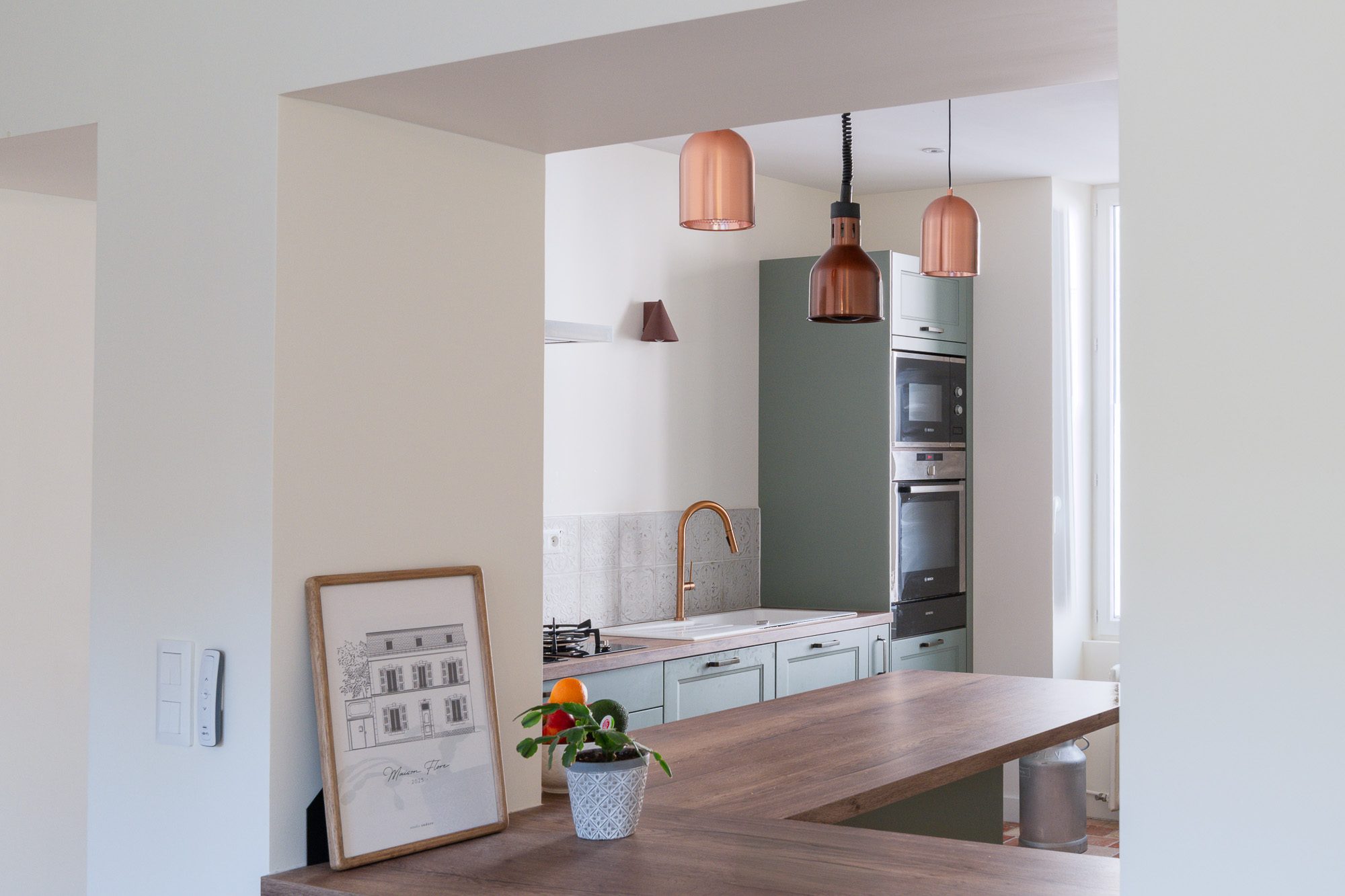 Cuisine ouverte vert sauge avec suspensions cuivrées et comptoir en bois – Projet Flore, rénovation d’une maison ancienne au Mans