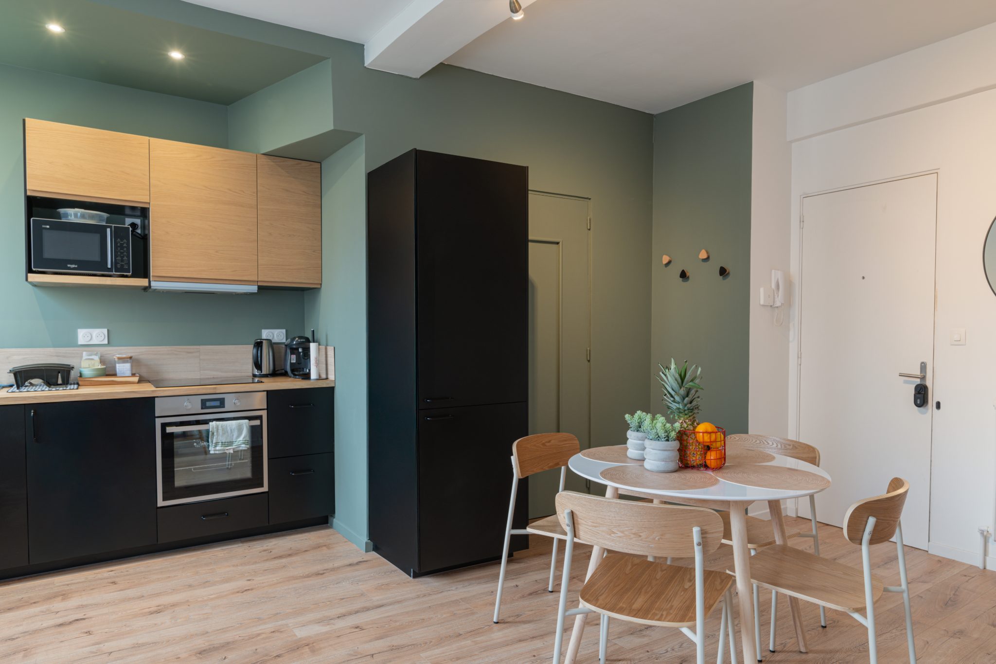 Pièce de vie d’un T2 rénové : cuisine noire et chêne, murs vert de gris et table ronde – Projet Ormeau au Mans