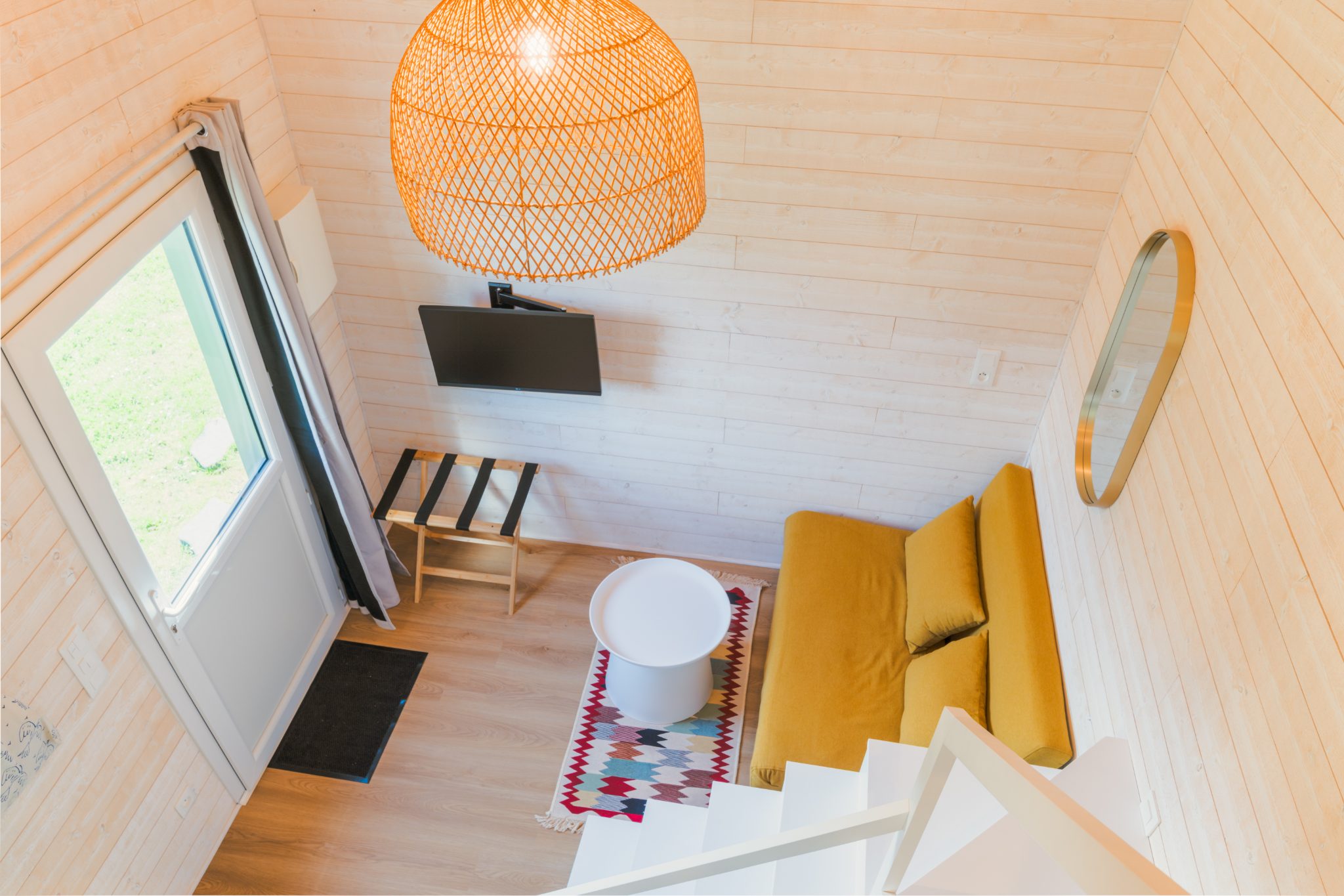 Vue plongeante sur le séjour d’une tiny house : banquette moutarde, suspension en rotin et lambris bois clair – Projet Tiny House en Sarthe